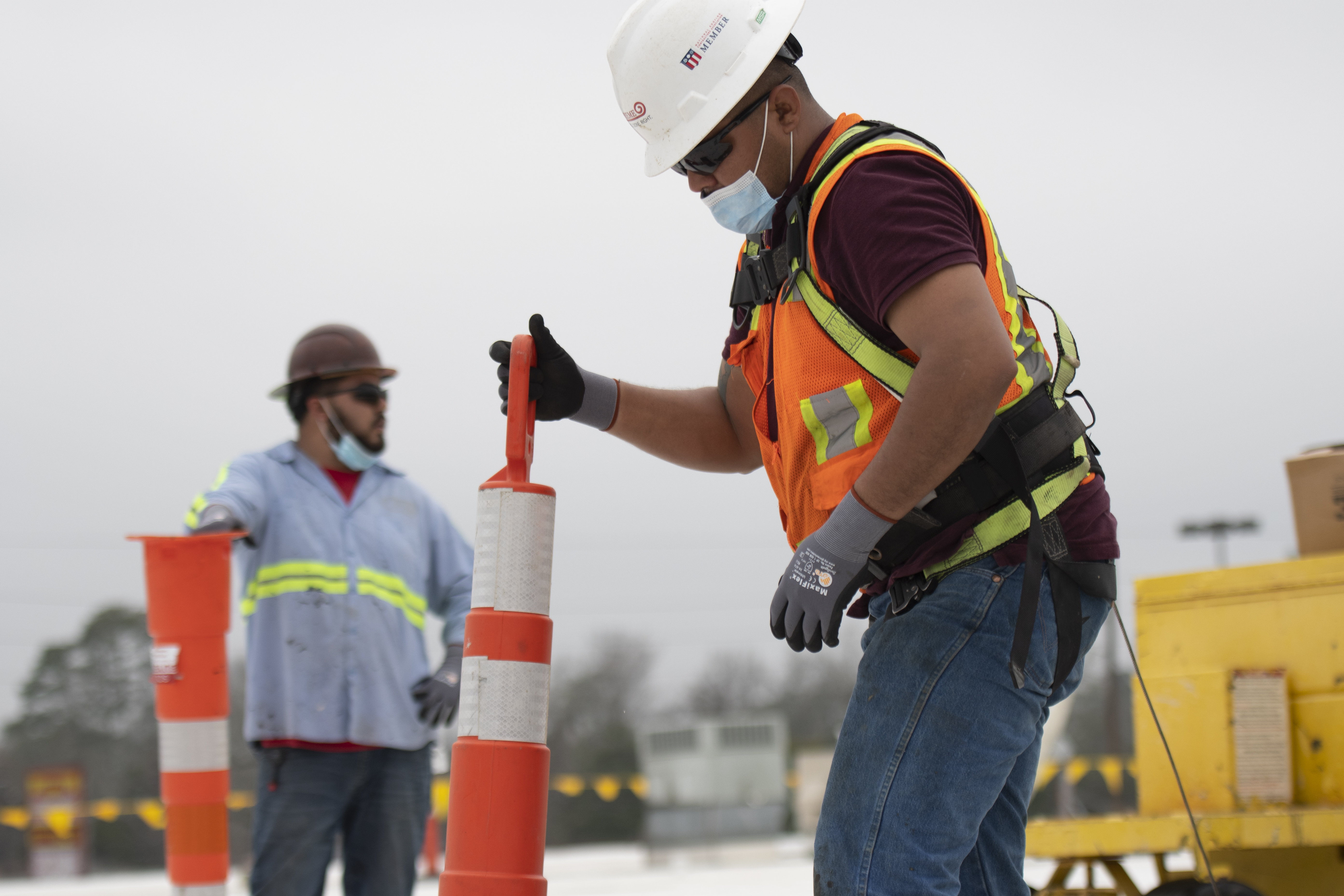 Construction worker wearing full safety equipment, including hard hat, gloves, reflective vest, face covering, and fall protection harness while placing traffic safety cones at an active job site.